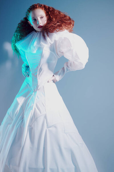 Sophisticated female model with lush red curly hair posing in a white art dress. A studio portrait with mixed red color lighting. Fashion art. 