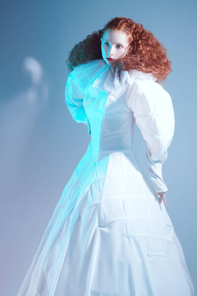 Beautiful female model with lush red curly hair posing in a white art dress on blue background. A studio portrait with mixed red color lighting in haze. Fashion art. 