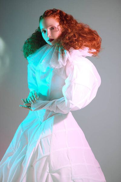 Redhead female model with lush curly hair posing in a white art dress on grey background. A studio portrait with green and red color lighting. Fashion art. 