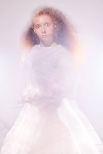 Portrait of a refined female model with lush red curly hair posing in a long white art dress among flashes and haze. Art fashion. 