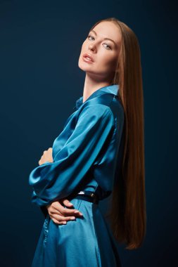 Moda fotoğrafı. Uzun düz saçlı güzel manken kız koyu mavi arka planda mavi saten elbiseyle poz veriyor.. 