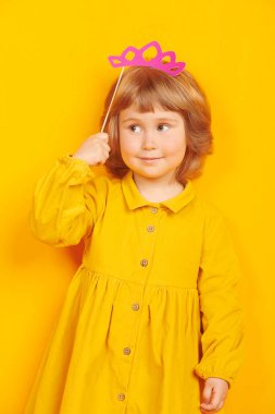 Prenses tacıyla oynayan ve gülümseyen tatlı küçük bir kız. Mutlu bir çocukluk. Çocukların duyguları ve hayal gücü. Sarı arka planda stüdyo resmi. 