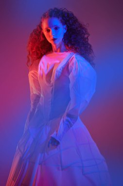 Beyaz bir sanat elbisesi içinde poz veren gür kırmızı kıvırcık saçlı sofistike bir kadın modelin portresi. Karanlık bir odada koyu kırmızı ışıklı bir stüdyo portresi. Moda sanatı. 