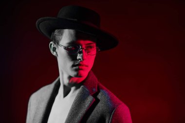 Paltolu, şapkalı ve gözlüklü kırmızı gri ışıklı yakışıklı bir manken. Erkekler için optik. Erkek modası. Portreyi kapat.