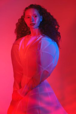 İnce, kırmızı kıvırcık saçlı, uzun beyaz fütüristik elbisesi ve neon kırmızısı ışıklandırmasıyla poz veren zarif bir kadın modelinin sanat portresi..
