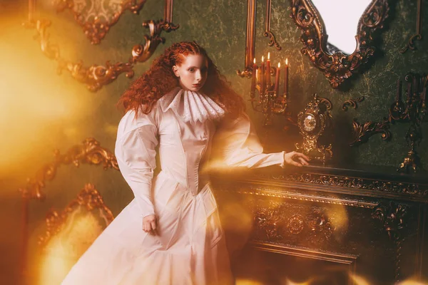 Portrait of a s sophisticated lady with lush red hair with fine curls standing in a vintage interior in art dress with a ruffled renaissance collar. History of fashion and hairstyles.