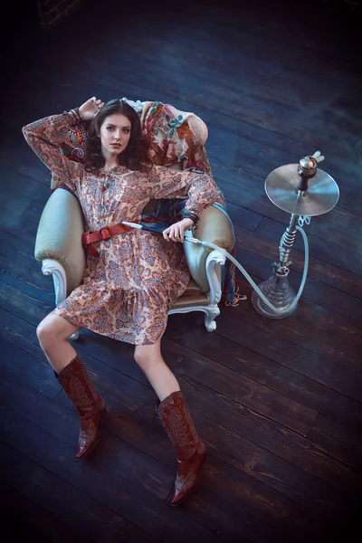 Bohemian style. Fashion shot of a beautiful young woman in boho style dress and jewelry posing in a vintage armchair. Full length portrait.