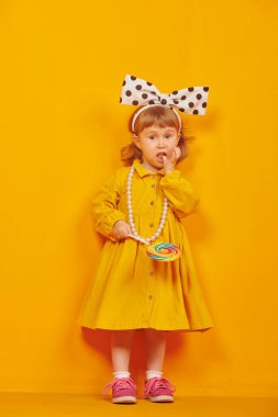 Şık sarı elbiseli sevimli küçük bir kız çocuğu ve parlak sarı arka planda büyük bir lolipopla büyük bir fiyonk. Çocuk modası. Tatlılar. 