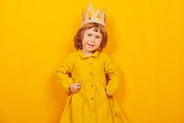 Kartondan taç giymiş şirin küçük bir kız kameraya bakıyor ve gülümsüyor. Mutlu bir çocukluk. Çocukların duyguları ve hayal gücü. Sarı arka planda stüdyo resmi. 