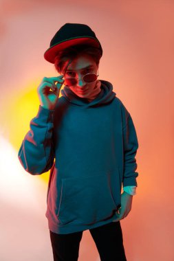 Modern genç çocuk kapüşonlu, kot pantolonlu, beyzbol şapkalı ve karışık renkli gözlüklü poz veriyor. Gençlik modası.