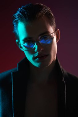 Genç bir adamın şık gözlüklü bir ceketle poz verirken yakın plan portresi. Karanlıkta çok renkli ışıklarda moda portresi. Erkekler için optik.