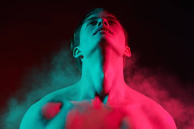 Siyah dumanlı arka planda çok renkli ışıklara bakan üstsüz bir gencin portresi. İnsanın güzelliği ve sağlığı. Resim..