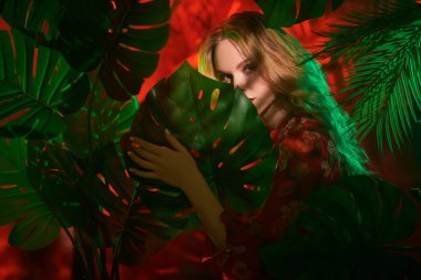 Uzun dalgalı sarı saçlı, parlak makyajlı, tropik yeşil yaprakların arasında kırmızı elbiseli güzel bir kadın model. Güzellik, moda. Orman fotoshoot. 