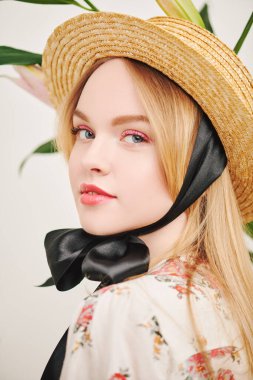 Hasır şapkalı güzel bir kız ve çiçekli beyaz bir arka planda poz veren bir yaz bluzu. Bahar ve yazın ilham kaynağı. Pembe renkli açık renk makyaj. Parfüm ve kozmetik.