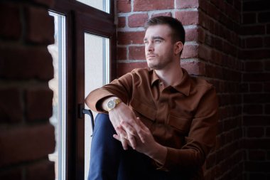 Sıradan giyinmiş yakışıklı bir adam pencerenin kenarında otururken pencereden dışarı bakar. Erkek güzelliği, moda. Sıradan stil.