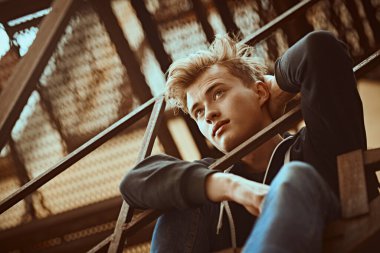 Sıradan giyinmiş yakışıklı genç adam metal endüstriyel bir merdivende oturuyor. Şehirli grunge stili. Erkek modası.