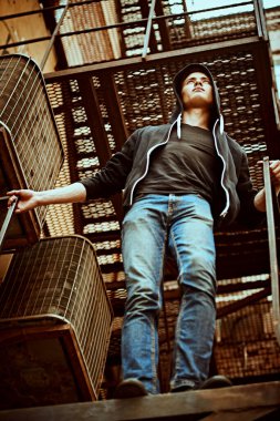 Günlük giysiler içindeki yakışıklı genç adam eski demir bir merdivende duruyor. Kentsel grunge tarzı.