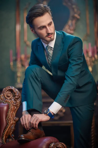 Handsome young man in elegant classic suit poses by a vintage armchair in a luxury apartment. Men's beauty, fashion. Luxury lifestyle.