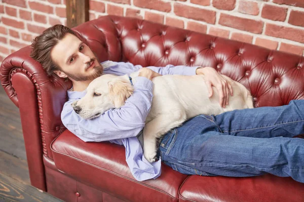 Happy calm young man lies on the couch and hugs his pet Golden Retriever dog.