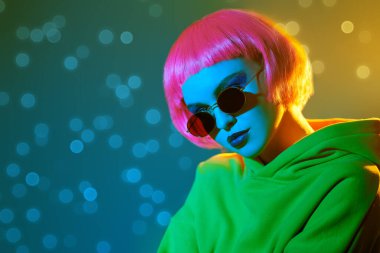 Parlak makyajlı, pembe saçlı, kırmızı güneş gözlüklü, sarı takım elbiseli, karışık renkli ışık saçan modern bir kız. Sportif şıklıkta. Gençlik modası ve güzellik eğilimleri. 