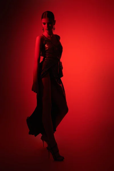Graceful young woman dancer poses in elegant long dress and high heels. Studio portrait in red light.