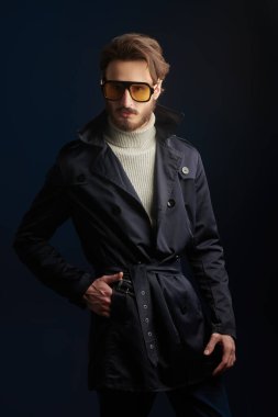 Siyah arka planda güneş gözlüğü ve yağmurlukla stüdyoda poz veren yakışıklı bir mankenin portresi. Erkek modası. 