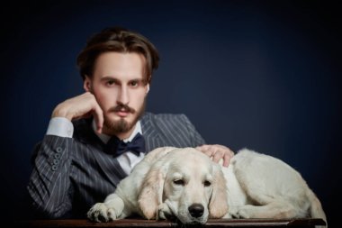 Moda, insan ve köpek. Zarif takım elbiseli yakışıklı bir adamın portresi. Siyah bir arka planda Golden Retriever köpeğiyle poz veriyor.. 