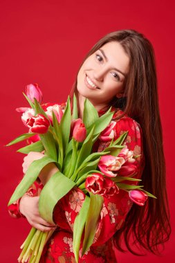 Bahar havası. Kırmızı çiçekli elbiseli güzel kız elinde bir buket lale çiçeği ve gülümseme tutuyor. Anneler Günü. Sevgililer Günü. Kırmızı arkaplanda stüdyo portresi.
