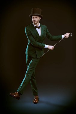 Men's fashion. Elegant gentleman in suit and top hat dances with a cane. Studio portrait on a dark blue background.  clipart