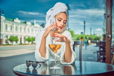 Kafasında beyaz bir havluyla beyaz geceliği olan çekici bir kız bir sokak kafeteryasında bir bardak şampanya ve telefonda dedikodularla oturuyor. Moda fotoğrafı.. 