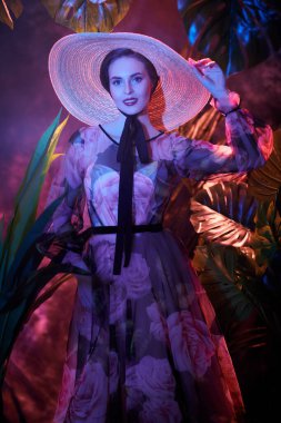 Açık çiçekli elbisesi ve geniş kenarlı hasır şapkası olan çekici yaz kızı tropik yeşillikler arasında poz veriyor. Yaz güzelliği, moda.