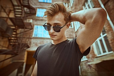 Gündelik kıyafetler ve güneş gözlüğü takan pozitif genç bir adam sokakta durup gülümsüyor. Şehirli grunge stili. Erkek modası.