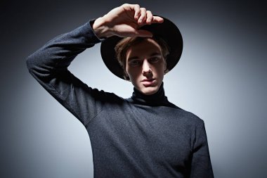 Geniş kenarlı şapkalı sanatçı genç adam ve balıkçı yaka kazak kameraya bakıyor. Gri arkaplanda stüdyo portresi.