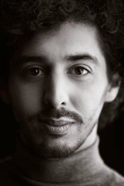 Kameraya bakıp gülümseyen kıvırcık saçlı yakışıklı bir gencin siyah beyaz portresi. Siyah arka plan. Erkek sağlığı ve güzelliği.