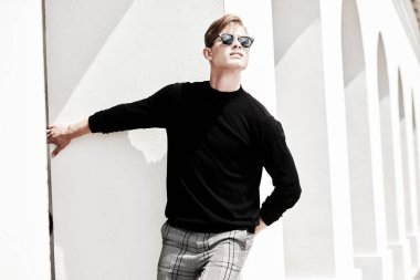 Moda fotoğrafı. Siyah kazaklı ve siyah güneş gözlüklü yakışıklı genç adam eski kasabadaki beyaz sütunların yanında poz veriyor. Erkek stili, güzellik. Resim..