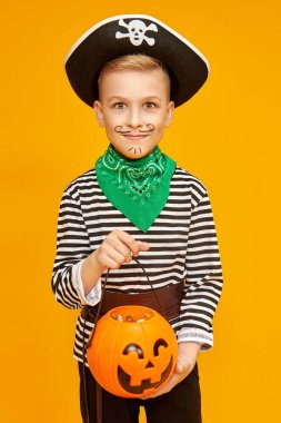 Mutlu Cadılar Bayramı! Stüdyoda sarı bir arka planda balkabağı sepeti ile poz veren korsan kostümlü neşeli çocuk.. 