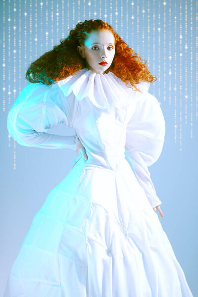 Fashion art history. Sophisticated female model with lush red curly hair posing in a white art dress with late renaissance ruffled collar. Studio portrait.
