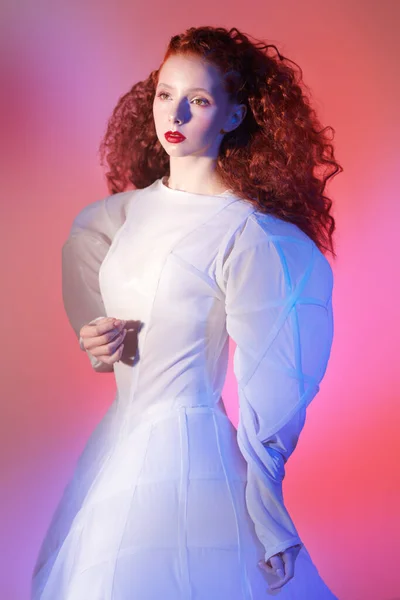 Fashion art. Portrait of a sophisticated female model with lush red curly hair posing in a white art dress. Studio portrait in mixed red color light.