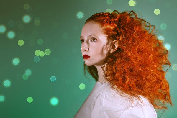 Hairstyling and makeup. Portrait of a refined fashion model girl with lush red curly hair posing in a white haute couture dress on a green background with lights. 