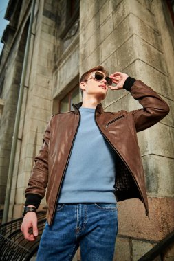 Güneş gözlüklü, deri ceketli, kendine güvenen genç bir adam eski bir binanın arka planında duruyor. Erkek modası. Yaşam biçimi.