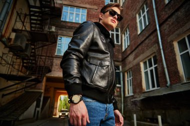 Siyah deri ceketli ve siyah güneş gözlüklü yakışıklı adam şehrin eski semtinde caddede duruyor. Kentsel grunge tarzı. 