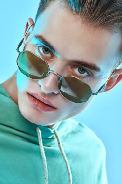 Moda eşofmanlı ve güneş gözlüklü yakışıklı bir gencin mavi arka planda kameraya bakarken yakın plan portresi. Gözlük modası. Stüdyo resmi.