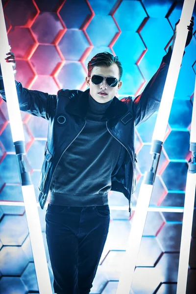 Men's style and fashion. Portrait of a courageous handsome man in black sunglasses and black leather jacket posing among neon lamps. Futurism, techno style.