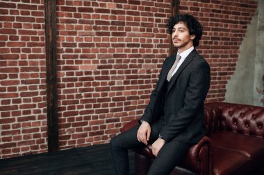 İş adamı portresi. Zarif klasik takım elbiseli yakışıklı esmer adam, dairesinin içindeki deri koltukta oturuyor. Erkek modası. 