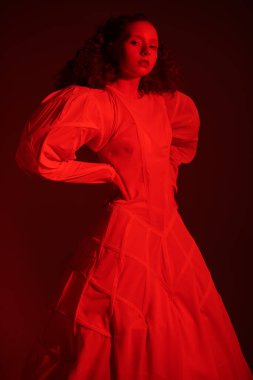 Kırmızı ışıkta beyaz bir sanat elbisesi içinde poz veren gür kırmızı kıvırcık saçlı sofistike bir kadın modelin portresi. Moda sanatı. Stüdyo çekimi. 