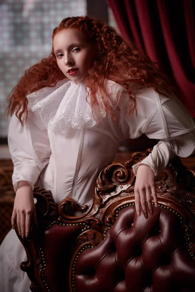 History of fashion and hairstyles. A sophisticated fashion model girl with lush red hair with fine curls poses in a vintage interior in art dress with a ruffled renaissance collar.