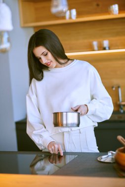 Mutlu esmer genç bayan mutfakta bir tencereyle yemek hazırlar ve gülümser. Sağlıklı beslenme ve beslenme. Modern iç mekan, mutfak mobilyası..