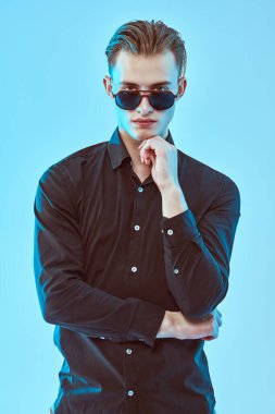 Mavi arka planda siyah gömlekli ve şık güneş gözlüklü yakışıklı bir genç adam portresi. Erkek modası. Aksesuarlar, optikler. 