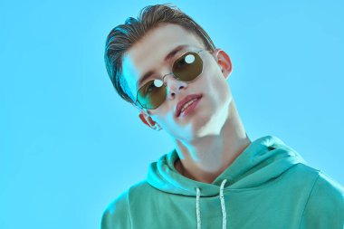 Yakışıklı genç adam modayla poz veriyor ve mavi arka planda şık güneş gözlüğü takıyor. Gözlük modası. Stüdyo resmi.
