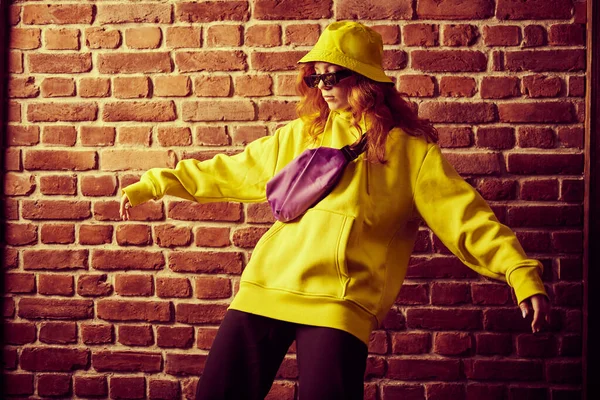 Street dance girl dancing in bright colorful clothes and sunglasses by a brick wall. Modern dances style.